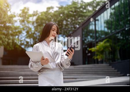 Eine wunderschöne, erfolgreiche asiatische Geschäftsfrau in stilvoller Bürokleidung, die einen Aktenordner hält, liest Nachrichten auf ihrem Smartphone, während sie hinuntergeht Stockfoto