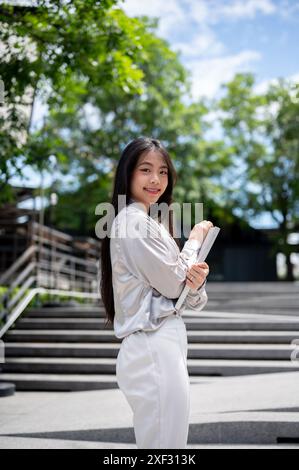 Eine schöne, glückliche asiatische Geschäftsfrau mit Aktenordner lächelt an einem sonnigen Tag draußen in der Stadt in die Kamera. Stockfoto