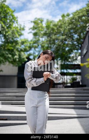 Eine beschäftigte, frustrierte asiatische Geschäftsfrau mit einem Aktenordner telefoniert mit ihrem Kollegen und überprüft die Zeit auf ihrer Armbanduhr Stockfoto