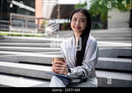 Eine schöne, erfolgreiche asiatische Geschäftsfrau in stilvoller Bürokleidung sitzt draußen auf der Treppe mit einem Kaffee zum Mitnehmen in der Hand und lächelt an Stockfoto
