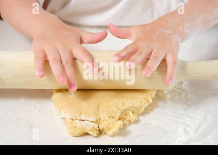 Ein kleines Kind wälzt auf einem Tisch Teig aus. Der Teig besteht aus Mehl und Butter und wird mit einer Nudelnadel flach gepresst. Das Kind ist fokussiert o Stockfoto