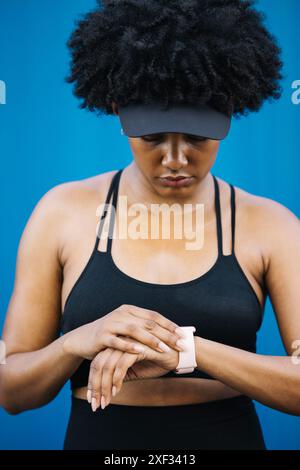 Porträt einer jungen, schönen Frau, die während eines Workouts ihre Smartwatch ansieht, während sie vor blauem Hintergrund steht. Stockfoto