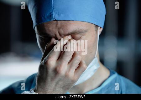 Nahporträt eines erschöpften Chirurgen, der die Augen im dunklen Operationssaal reibt Stockfoto