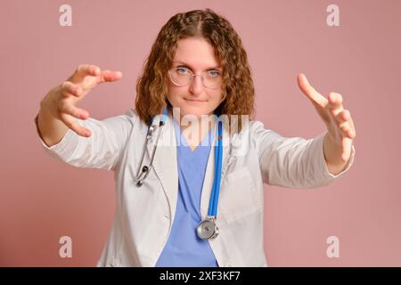 Eine Ärztin, die mit den Händen zu einem Patienten streckt, rosa Hintergrund des Studios. Krankenschwester in Uniform mit Stethoskop auf rotem Atelierhintergrund Stockfoto