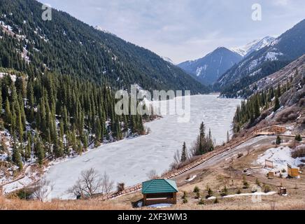 Malerischer, eisbedeckter Kolsai-See in Kasachstan, umgeben von Hügeln, die mit grünen Fichten und Schnee bedeckt sind. Fußgängerzone, hölzerner Öko-Trail um den herum Stockfoto