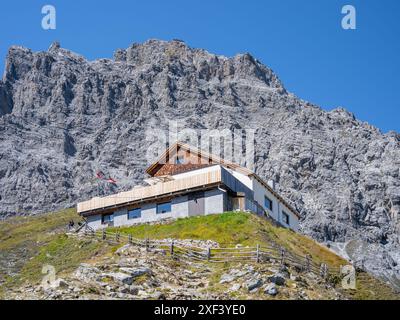 Eine traditionelle Almhütte, die Tabarettahütte, liegt an einem üppig grünen Hang unter dem imposanten Ortlerberg unter einem klaren blauen Himmel. Stockfoto