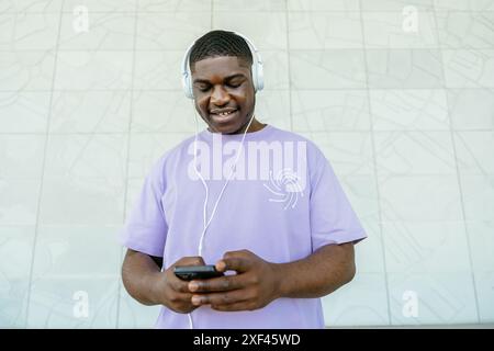 Ein junger Mann hört Musik auf seinen Kopfhörern, während er ein Handy hält. Er lächelt und amüsiert sich Stockfoto