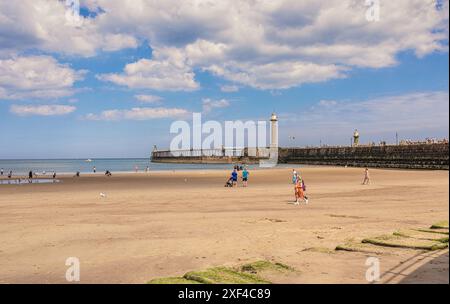 Ein Pier mit einem Leuchtturm erstreckt sich an einem hellen sonnigen Tag bis zum Meer. Im Vordergrund steht ein Strand, an dem Urlauber den Sand genießen. A Sky wi Stockfoto