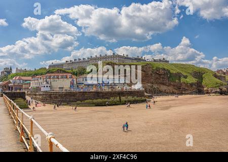 Ein Strand an einem hellen, sonnigen Tag mit Urlaubern, die den Sand genießen. Es gibt eine Ufermauer mit Fahrgeschäften und einen Hügel mit Hotels oben. Geländer sind Stockfoto