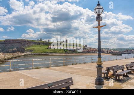 Blick auf einen weit entfernten Hügel von einem Pier mit Straßenlaterne und Bänken. Häuser schmiegen sich unter der Klippe mit einer Kirche und Whitby Abbey auf dem Hügel. A Stockfoto