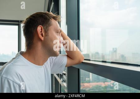 Unglücklicher frustrierter Mann, der an Depressionen und schlechten Nachrichten litt und auf ein Fenster mit Blick auf die Stadt blickt. Müder, trauriger Mann zu Hause. Psychische Gesundheit, Lebensprobleme Stockfoto