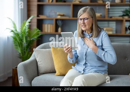 Ältere Frau hat Brustschmerzen, während sie Tablette verwendet, um einen Arzt online zu konsultieren. Sie sitzt auf einem Sofa in einem stilvollen Wohnzimmer, sieht besorgt aus und hält ihre Brust. Stockfoto