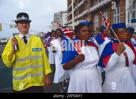 Die Church of God of Prophecy, die Women's Missionary Band, steht an einem Sonntagmorgen vor dem Brighton Convention Centre für ihre jährliche National Convention, die mit einer Parade am Meer beginnt. Sie tragen eine Kirchenfahne, die das Zepter zeigt, um Christi Autorität zu symbolisieren, einen Stern, der den Stern von Bethlehem und die Krone des Königtums darstellt. Die Farbe Violett symbolisiert Könige, weiß und blau für die Wahrheit, und Rot steht für das Blut Jesu. Eine Polizistin geht mit ihnen. Brighton, East Sussex, England um die 1985 1980er Jahre Großbritannien HOMER SYKES Stockfoto