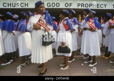 Die Church of God of Prophecy, die Women's Missionary Band, steht an einem Sonntagmorgen vor dem Brighton Convention Centre für ihre jährliche National Convention, die mit einer Parade am Meer beginnt. Sie tragen eine Kirchenfahne, die das Zepter zeigt, um Christi Autorität zu symbolisieren, einen Stern, der den Stern von Bethlehem und die Krone des Königtums darstellt. Die Farbe Violett symbolisiert Könige, weiß und blau für die Wahrheit, und Rot steht für das Blut Jesu. Brighton, East Sussex, England um die 1985 1980er Jahre Großbritannien HOMER SYKES Stockfoto