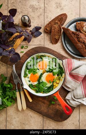 Grüne Shakshuka. Spiegeleier, Kartoffeln, Spinat und Zwiebeln in roter gusseiserner Pfanne mit Roggenbrot auf alten Fliesen Tischhintergrund. Traditionell Jüdisch, Israel Stockfoto