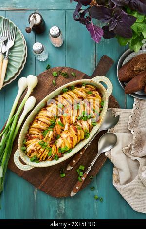 Hasselback-Kartoffel gefüllt mit Schinken, Zwiebeln, Knoblauch und Kräutern, serviert in ovaler Keramikform mit Gewürzen, Basilikum, Koriander, Roggenbrotstücken auf Blau Stockfoto