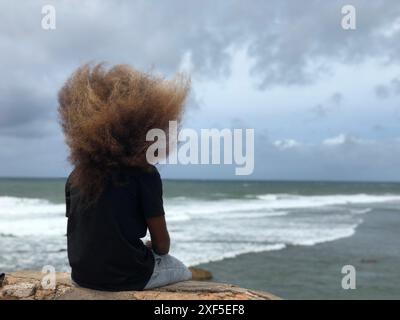 Ein einsamer Mann mit langen Haaren, der an einem windigen Tag am Strand sitzt Stockfoto