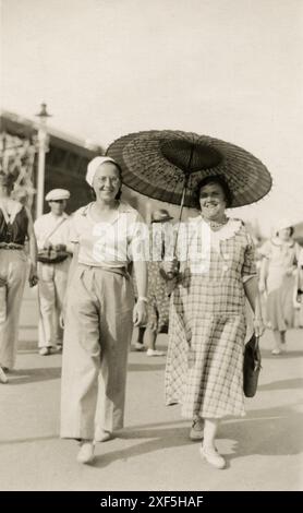 Margate, Kent. 1930er Jahre – Ein Straßenfoto von zwei Frauen, die entlang der Promenade in Margate, Kent, England, spazieren gehen. Die ältere Frau trägt ein kariertes Baumwollkleid und trägt einen Sonnenschirm im chinesischen Stil. Die jüngere Frau trägt ein T-Shirt mit Kragen, Hosen und einer Matrosenmütze. Beide Frauen lächeln. Stockfoto