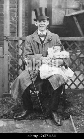 England. Ca. 1910er Jahre – Ein stolzer junger Vater saß draußen und hielt sein Baby in einem Taufmantel. Der Mann ist elegant gekleidet, trägt einen Hut, einen Mantel mit einem floralen Knopflochdekor und eine Schleife mit einer Goldnadel. Ein Gehstock mit silberner Spitze ruht an seinem Knie. Der Mann lächelt, seine Zähne zeigen Anzeichen von Karies. Stockfoto