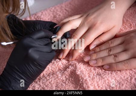 Kosmetikerin, die Nagelhaut von Klientennägeln mit einem Spezialwerkzeug in einem Kosmetikstudio entfernt. Die Kosmetikerin trägt Schutzhandschuhe Stockfoto