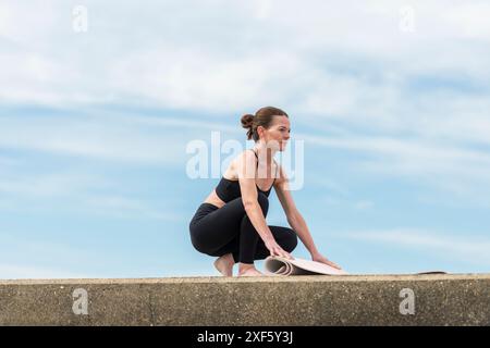 Frau, die ihre Yoga-Matte ausrollt, draußen mit blauem Himmel Hintergrund Stockfoto