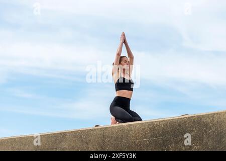 Sportliche Frau macht Yoga und Entspannungsübungen auf einer Matte draußen, blauer Himmel Hintergrund Stockfoto