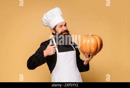 Lächelnder männlicher Koch in Uniform mit Kürbis, der Daumen nach oben zeigt. Herbstrezepte. Saisonale Vitamine. Gesundes Bio-Erntegemüse. Bärtiger Mann im Koch Stockfoto