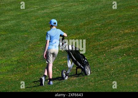 Der junge Teenager wartet während eines Golfturniers auf ihn. Er steht, beiläufig neben seinen Golfschlägern. Er trägt ein blaues Hemd und eine blaue Mütze. Stockfoto