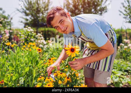 Glücklicher junger Mann, der Blumen im Sommergarten pflückt. Gärtner genießt gelbe Blüten mit Schürze und Blick in die Kamera. Freizeitaktivitäten im Freien Stockfoto