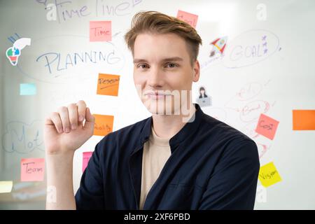 Porträt eines glücklichen jungen Geschäftsmannes in blauer, lässiger Uniform, während er vor einem Glasbrett mit bunten Haftnotizen und Mindmap mit Co steht Stockfoto