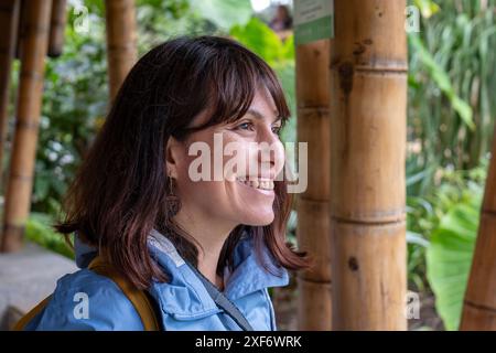 Eine junge braune Frau in einem blauen Mantel mit einem Kameraband um den Hals lächelt und starrt an Einem regnerischen Tag den Himmel an, mit Bambusstangen im Hintergrund Stockfoto