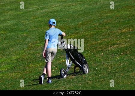 Der junge Teenager wartet während eines Golfturniers auf ihn. Er steht, beiläufig neben seinen Golfschlägern. Er trägt ein blaues Hemd und eine blaue Mütze. Stockfoto