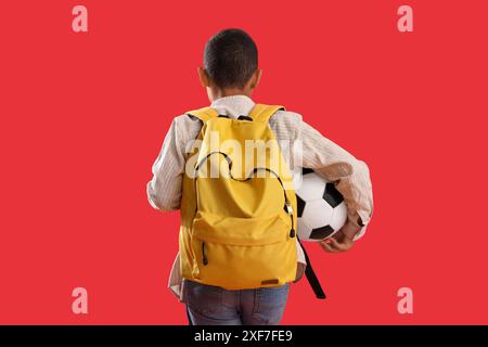 Kleiner Schuljunge mit Rucksack und Fußball auf rotem Hintergrund, Rückansicht Stockfoto