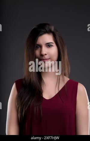 Eine junge Frau mit langen braunen Haaren posiert in einem Studio-Setting, trägt ein burgunderrotes Kleid vor einem neutralen grauen Hintergrund Stockfoto