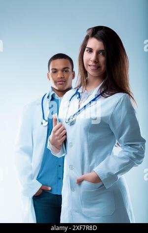 Zwei selbstbewusste Ärzte in weißen Mänteln und Stethoskopen posieren in einem Atelier. Sie repräsentieren verschiedene Rollen im Gesundheitswesen und betonen Teamarbeit und Vertrauen in Pat Stockfoto