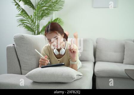 Glückliche junge asiatische Frau in legeren Kleidern, die auf der Couch liegt und ein digitales Tablet benutzt Stockfoto