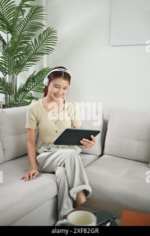 Porträt einer lächelnden jungen asiatischen Frau in schnurlosen Kopfhörern mit digitalem Tablet auf der Couch Stockfoto