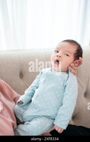 Ein entzückender, verschlafener neugeborener Sohn im süßen blauen Pyjama ist in der Umarmung seiner Mutter auf einer Couch im Wohnzimmer. Ein gähnender Säugling im Pyjama. Kind Stockfoto