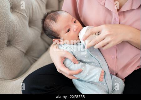 Ein entzückendes, verschlafenes Neugeborenes im blauen Pyjama ist in den Armen seiner Mutter und trinkt Milch aus einer Flasche. Säuglingsmilchfütterungskonzept Stockfoto