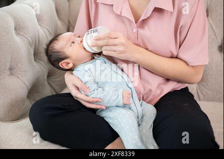 Ein entzückendes, verschlafenes Neugeborenes im blauen Pyjama ist in den Armen seiner Mutter und trinkt Milch aus einer Flasche. Säuglingsmilchfütterungskonzept Stockfoto