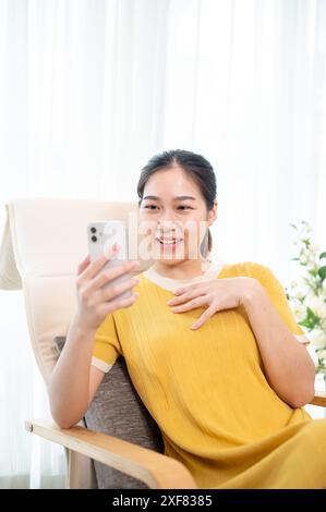 Eine glückliche asiatische Frau in einem gelben Kleid benutzt ihr Smartphone, während sie sich auf einem Sessel im Wohnzimmer entspannt. Auf dem Telefon scrollen, Nachrichten lesen, Stockfoto