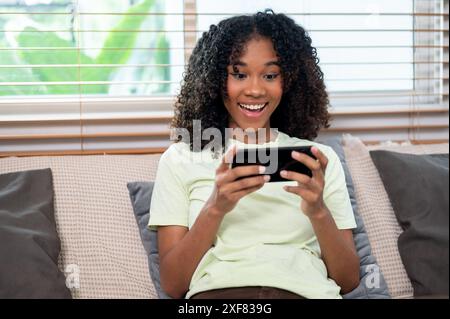 Eine fröhliche, junge schwarze Frau spielt gern ein mobiles Spiel oder sieht Videos auf ihrem Smartphone an, während sie sich auf einer Couch im Wohnzimmer entspannt, spe Stockfoto