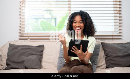 Eine junge, fröhliche schwarze Frau ist begeistert über etwas auf ihrem digitalen Tablet, das Tablet verwendet, einen Stift hält und zu Hause auf der Couch sitzt. Personen A Stockfoto