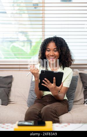 Eine junge, fröhliche schwarze Frau ist begeistert über etwas auf ihrem digitalen Tablet, das Tablet verwendet, einen Stift hält und zu Hause auf der Couch sitzt. Personen A Stockfoto