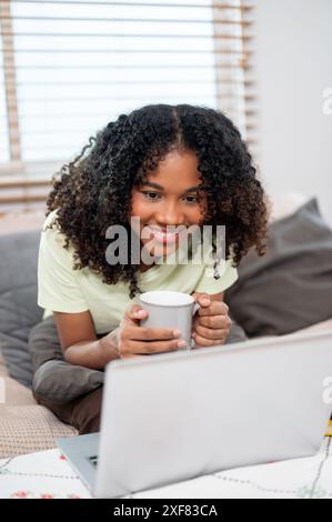 Eine attraktive, glückliche Schwarze liest gern Chats oder schaut sich einen Film auf ihrem Laptop an, während sie morgens auf der Couch zu Hause einen Kaffee trinkt Stockfoto