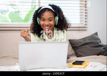 Eine fröhliche junge schwarze Frau mit Kopfhörern sitzt zu Hause auf einer Couch und zeigt ihre Faust, aufgeregt und überrascht von guten Nachrichten auf ihrem Laptop. Erhalten Stockfoto