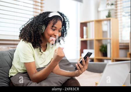 Eine fröhliche junge schwarze Frau mit Kopfhörern sitzt zu Hause auf einer Couch und zeigt ihre Faust, aufgeregt und überrascht von guten Nachrichten auf ihrem Smartphone. Stockfoto