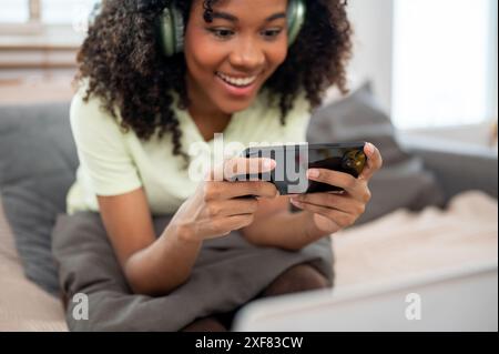Ein Nahbild einer fröhlichen, lustigen jungen Schwarzen Frau, die Kopfhörer trägt, spielt ein mobiles Spiel auf der Couch im Wohnzimmer. People und wi Stockfoto