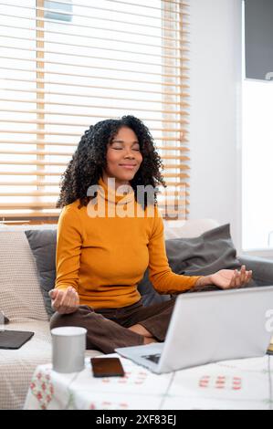 Eine ruhige, positive schwarze Frau meditiert auf einer Couch im Wohnzimmer, bevor sie mit der Arbeit beginnt. Achtsamkeit, positive Einstellung, Entspannung, Stresssituation Stockfoto