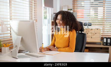 Eine attraktive, lächelnde schwarze Geschäftsfrau macht Notizen in einem Notizbuch, während sie Informationen auf ihrem Computerbildschirm ansieht und an ihrem Schreibtisch in t arbeitet Stockfoto